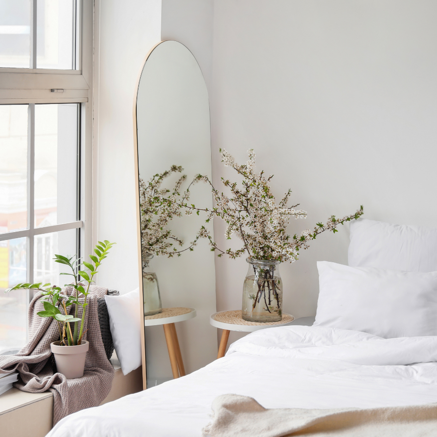 Serene bedroom with a white bed, large arched mirror, and vase of flowering branches on a round side table near the window.